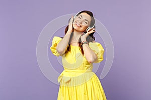 Smiling young woman in yellow dress keeping eyes closed, listen music, putting hands on headphones  on pastel
