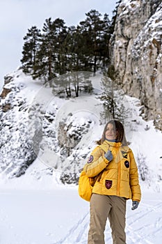 Smiling young woman in yellow jacket with backpack walking in winter by rocks covered snow, hiking in mountains outdoors activity