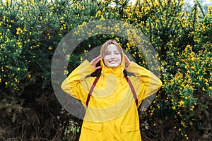 Smiling young woman in a yellow raincoat and hood enjoying walk on background of blooming bush with flowers. Travel