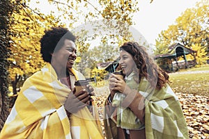 Smiling young women friends chatting outdoors and drink coffee while enjoying walk in park together