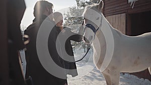 Smilling blond woman and tall bearded man standing with white horse at the snow winter ranch. Girl strokes animal. Happy