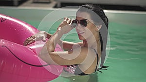 Smilling woman in sunglasses holding pink rubber ring in the water in swimming pool. Leisure of lonely lady in bikini.