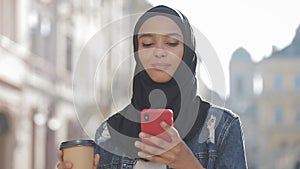 Smilng muslim woman wearing hijab headscarf walking in the city center, using smartphone and drinking coffee