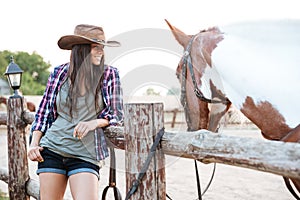 Smilng pretty young cowgirl standing with her horse on ranch