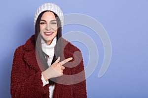 Smilng young brunette woman with long dark hair and red pomade,female dresses faux fur coat and white winter cap pointing aside