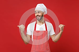 Smirked young bearded male chef cook baker man in striped apron white t-shirt toque chefs hat posing isolated on red