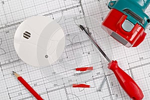 Smoke detector or fire alarm sensor on white architectural plans background with drill, screwdriver and screws, house safety or