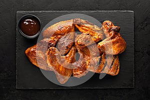 Smoked chicken wings on a slate board and barbecue sauce in a bowl on a rustic black background