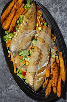 Smoked mackerel with vegetables and sweet potato fries on a black oval plate.