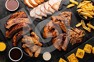 Smoked pork ribs and grilled chicken wings on a slate board, grilled corn and fries on the side on a black background.