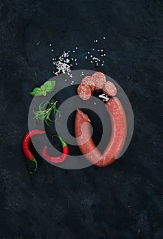 Smoked salami sausage with rosemary, chili pepper, basil and salt over black slate stone background, top view.
