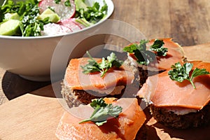 Smoked salmon canapes on rye bread with cream cheese and parsley on cut board and fresh vegetable salad in white bowl