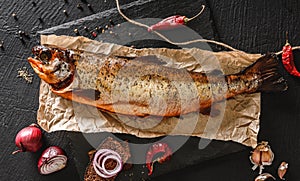 Smoked trout fish, red whole fish with spices and onion on craft paper over dark stone background. Seafood, top view