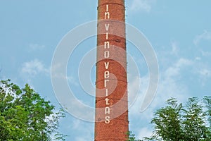 Smokestack with text In vino veritas. Industrial chimney on cloud sky background and tree crowns. Manufacturing of alcohol drinks