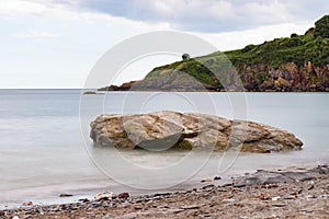 Smooth, calm sea around a shoreline rock at St Mary`s Bay beach in Torbay, Devon, with some coastline cliffs in the background