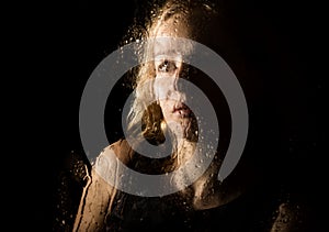 Smooth portrait of model, posing behind transparent glass covered by water drops. young melancholy and sad woman