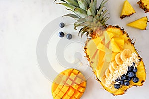 Smoothie bowl in a pineapple with coconut, bananas, mango & blueberries, top view on marble with copy space