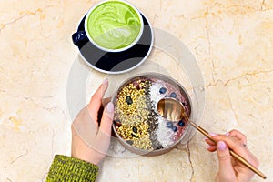 Smoothie bowl raw food, and matcha latte green tea cup, and female hands with metal spoon, ready to eat, on the table