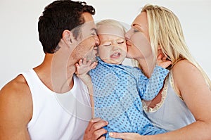 Smothering him with kisses. Two loving parents kissing their son on his cheeks while he looks upset and tries to push