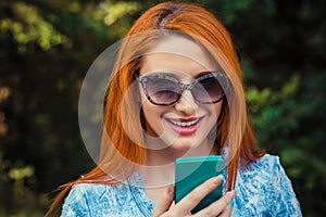 Sms. Young red head Woman in blue jeans texting looking talking to her Mobile Phone While Walking in a City park, green outdoors