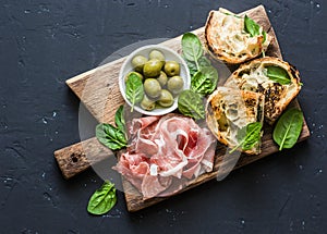 Snack board - prosciutto, olives, grilled mozzarella spinach sandwiches on dark background, top view. Mediterranean style snack, a