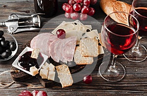 Snack with red wine, slices of cheese ,  ham, olives, cracker, jam, grapes, baguette,  corkscrew, on a dark wooden background, top