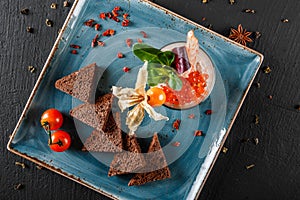 Snack from shrimps and red caviar with black bread, decorated with physalis and greens on plate over black background.Healthy food