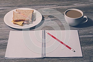 Snack at work. Tasty cheese sandwich on a plate, cup of hot coffee, open notebook with pencil on grey wooden background. Top view,