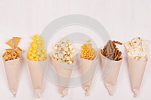 Snacks collection in craft paper cornets on white wooden board, top view, border.