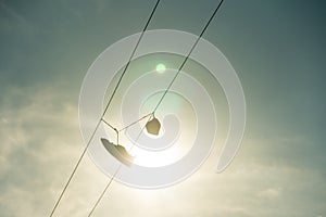 Sneakers hang on electric line wire cable with sun and sky on background. Funny urban outdoor footwear pair silhouette
