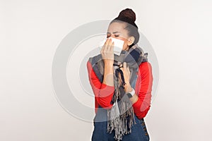 Sneeze and cough, flu symptoms. Portrait of unhealthy young woman wrapped in warm scarf cleaning nose with napkin