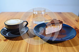 Snickers cheesecake on a blue saucer, a glass of water and a cup of fresh cappuccino on a large bright wooden table