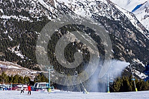 Snow canons blowing fresh snow on ski slope. Skiers on slopes with chairlifts and Pyrenees mountains in background.