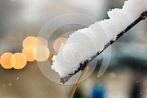 Snow cap of white fluffy snow on a branch of deciduous tree on a blurred background of cars with lights on in the city landscape