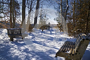 Snow-covered bench at a hiking trail with the silhouette of a man at the winter forest