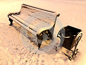 Snow covered bench and trash bin at night