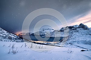 Snow covered mountain range with illuminated town on coastline in blizzard