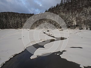 Snow covered partly frozen forest pond or lake with water stream creek with spruce tree forest, idyllic winter landscape