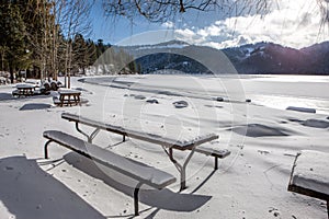 Snow covered picnic tables by frozen lake.