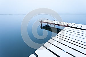 A snow-covered pier with a calm lake on a foggy day