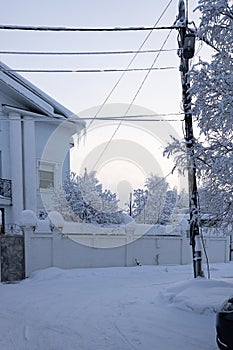 A snow-covered road, a leaning pole, a fence and the corner of a private residential house. A day during the polar night.