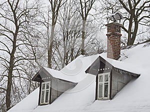 snow covered roof with brick chimney and bay bow window with icicles and bare tree background