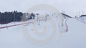 Snow-covered ski and snowboard pistes, equipped with a chair elevator and a rope tow elevator, mesh fence and lampposts, surrounde photo