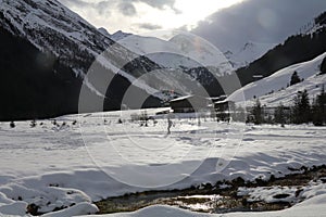 snow covered valley with a farmhouse and a single cross country skiier in the distance while the sun comes through the dark clouds