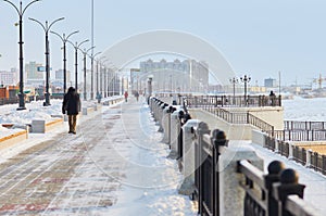 Snow-covered winter city embankment. People walk along the promenade. Lanterns, leaves, fences and drifts. Cold morning.