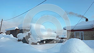 Snow covers a car parked in front of a house on a snowy slope