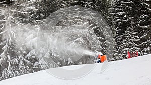 Snow gun spraying artificial ice crystals to ski piste, snowmaking in winter sports resort, trees in background