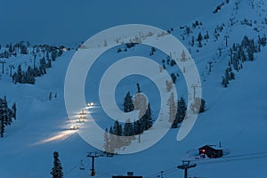 Snow machines with headlights on grooming ski slopes at twilight/dusk on a mountain with trees and a cabin