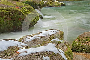 Snow on mossy rocks beside smooth cool flowing river water
