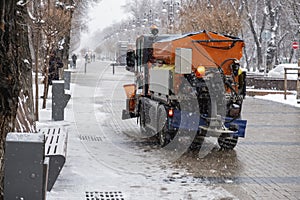 Snow plow is sprincling salt or de-icing chemicals on pavement in city. Cleaning service. Frost winter season.